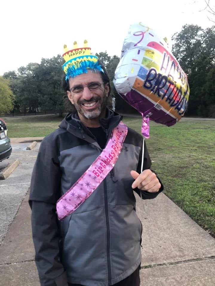 Larry outdoors wearing a birthday crown and sash and holding a birthday balloon