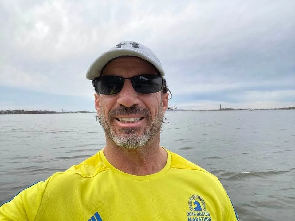 Larry taking a selfie on a run beside New York Harbor in his 2019 Boston Marathon shirt, the Statue of Liberty in the distance